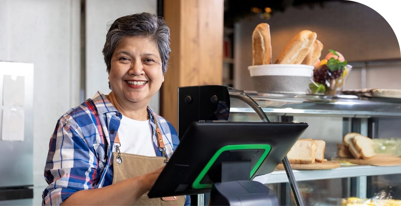 A smiling older woman in an apron manages a point of sale screen besides a case full of baked goods