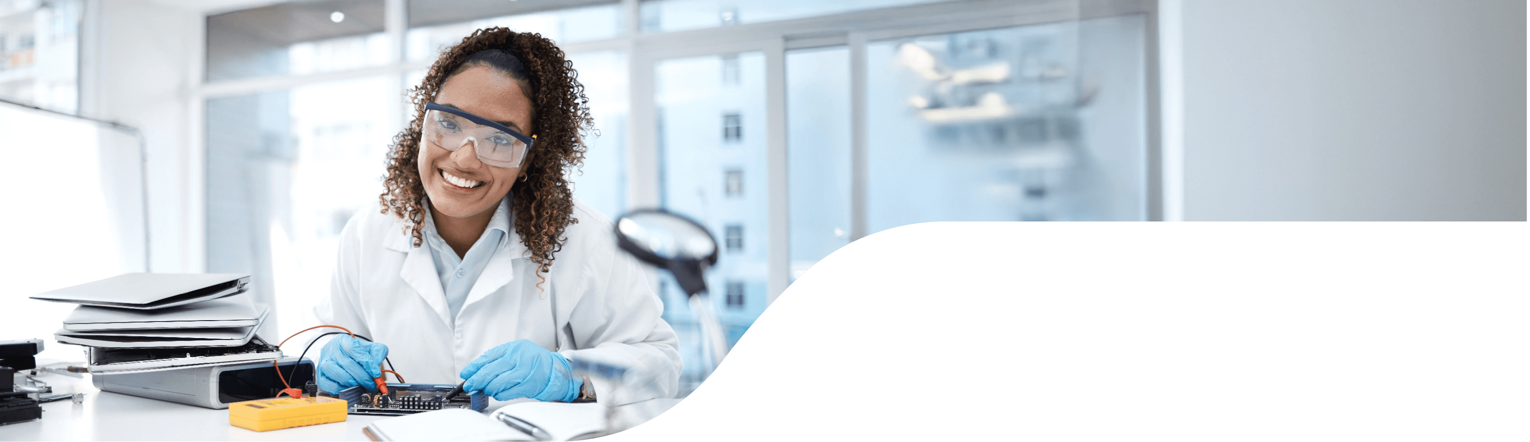 A smiling woman in a lab coat wearing safety glasses works on a circut borad with testing equipment