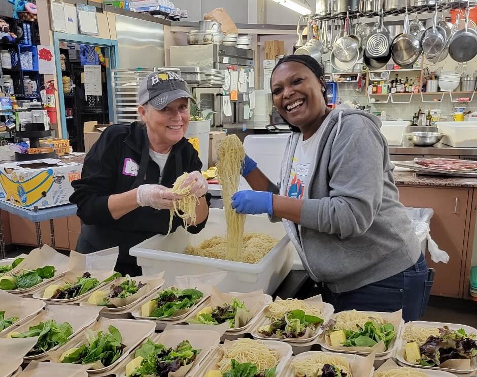 Two women are working and smiling while preparing spaghetti in a kitchen.