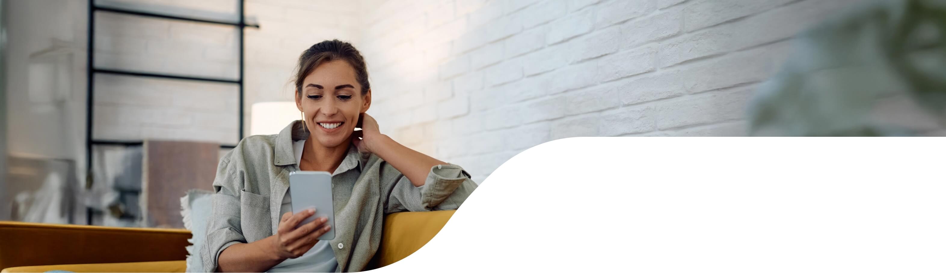 A smiling woman looking down at her smartphone. She's sitting on a couch within a modern looking space with exposed brick that is painted white.