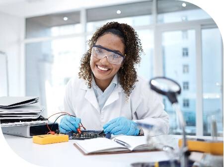 A smiling woman in a lab coat wearing safety glasses works on a circut borad with testing equipment