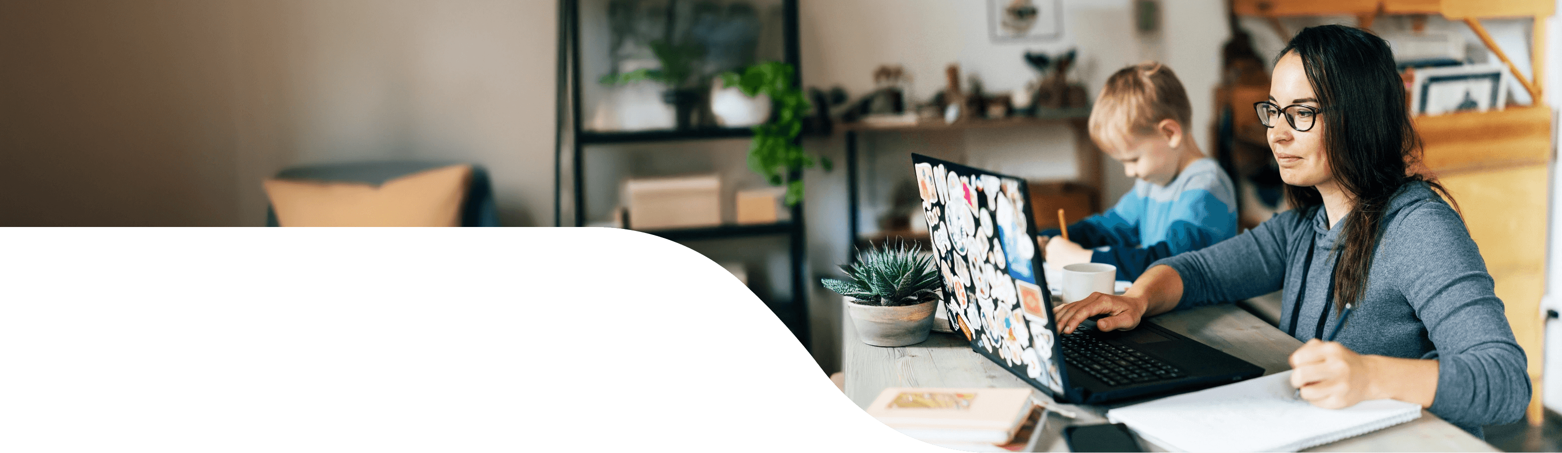 A woman with a young child in the background works at a laptop covered with stickers